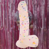 The Get Lucky Confetti Cock 8.5" Silicone Dildo, festooned with vibrant patterns, stands boldly against a gleaming pink tinsel backdrop, reminiscent of a whimsical party accessory in its cheeky presentation.